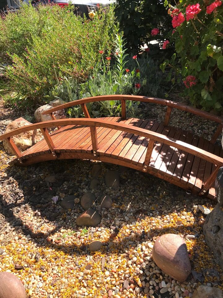 Arched timber garden bridge with curved handrails spanning a pebble creek bed among flowering shrubs