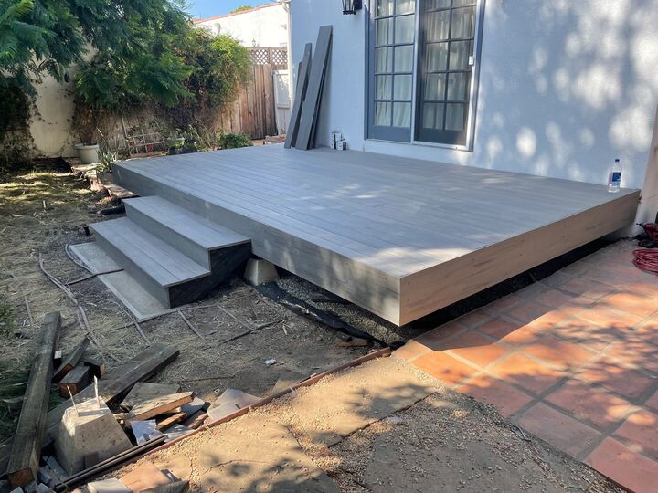 Freshly built grey deck platform and steps beside a white stucco wall with offcuts on the ground