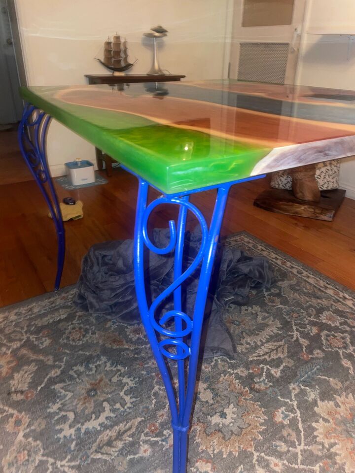 Green resin river table indoors on blue scrollwork legs, lit from a window