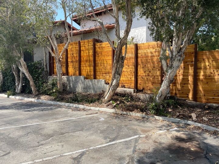 Long horizontal redwood fence running past mature trees at the edge of a parking area