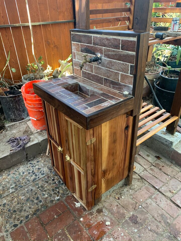 Outdoor timber sink cabinet with a tiled top and doors, standing on a brick patio