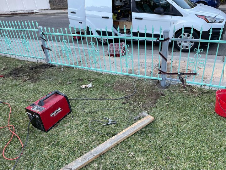 Sections of green wrought iron fence laid out on the grass beside a welding generator during the repair